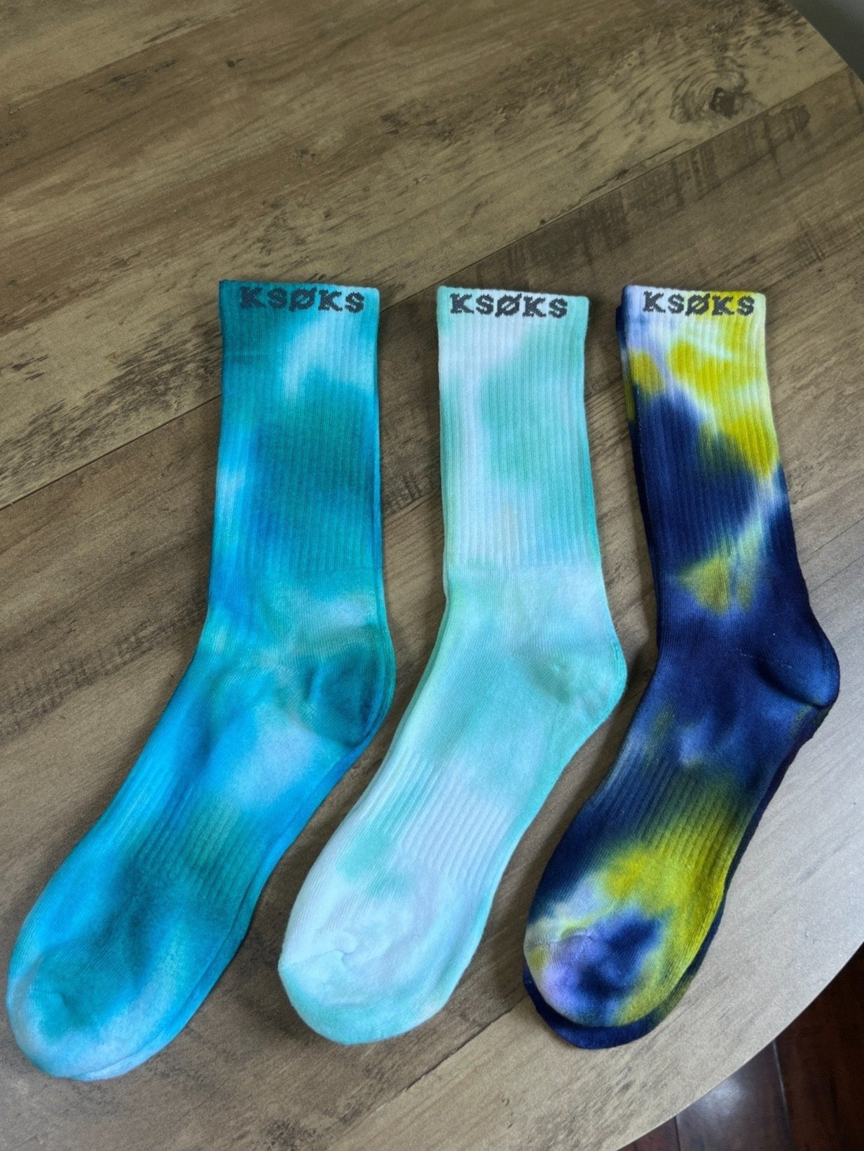 Three pairs of tie-dye socks on a wooden surface with 'KSOKS' branding.