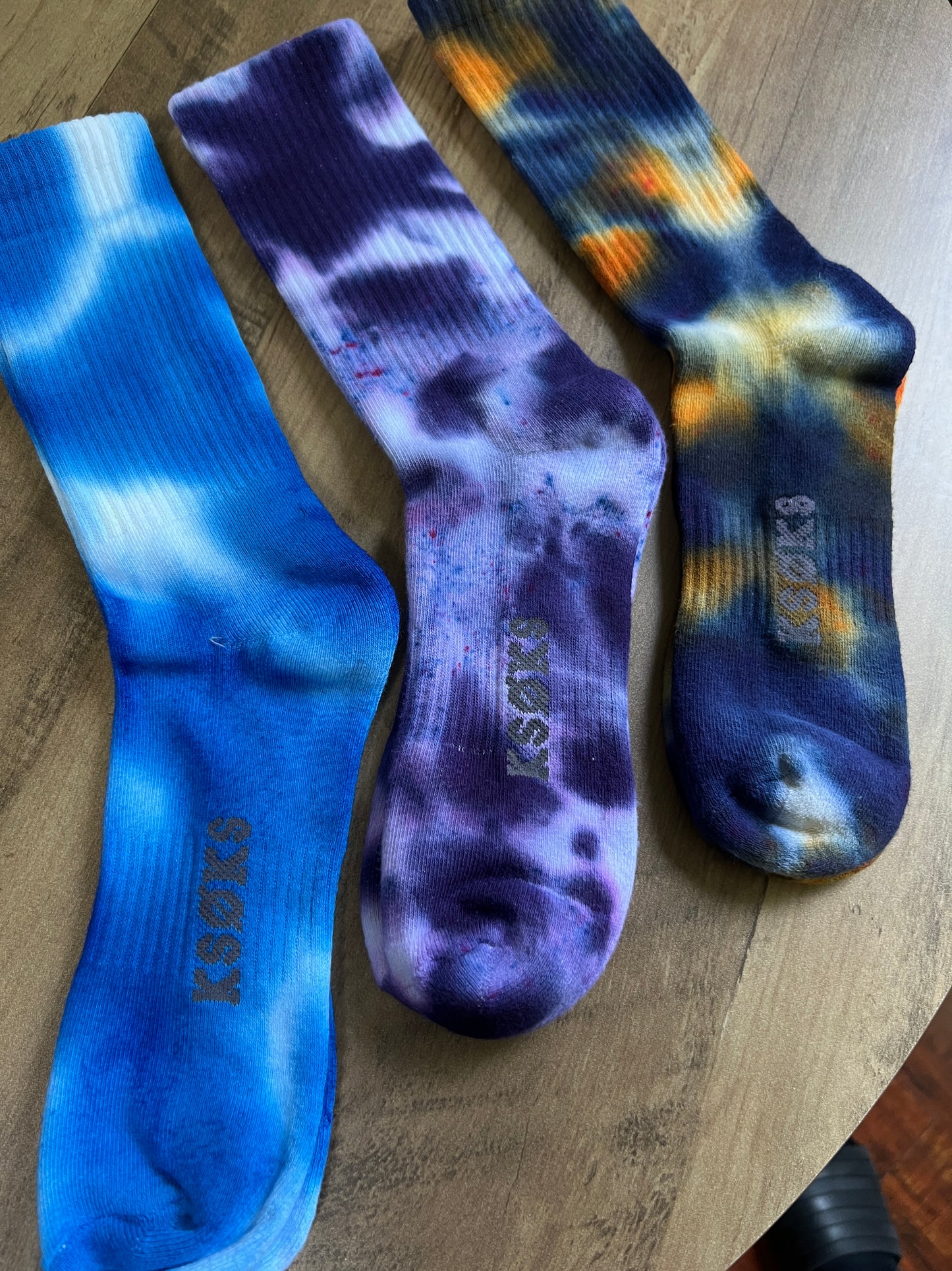 Three pairs of tie-dye socks with a brand logo on a wooden surface.
