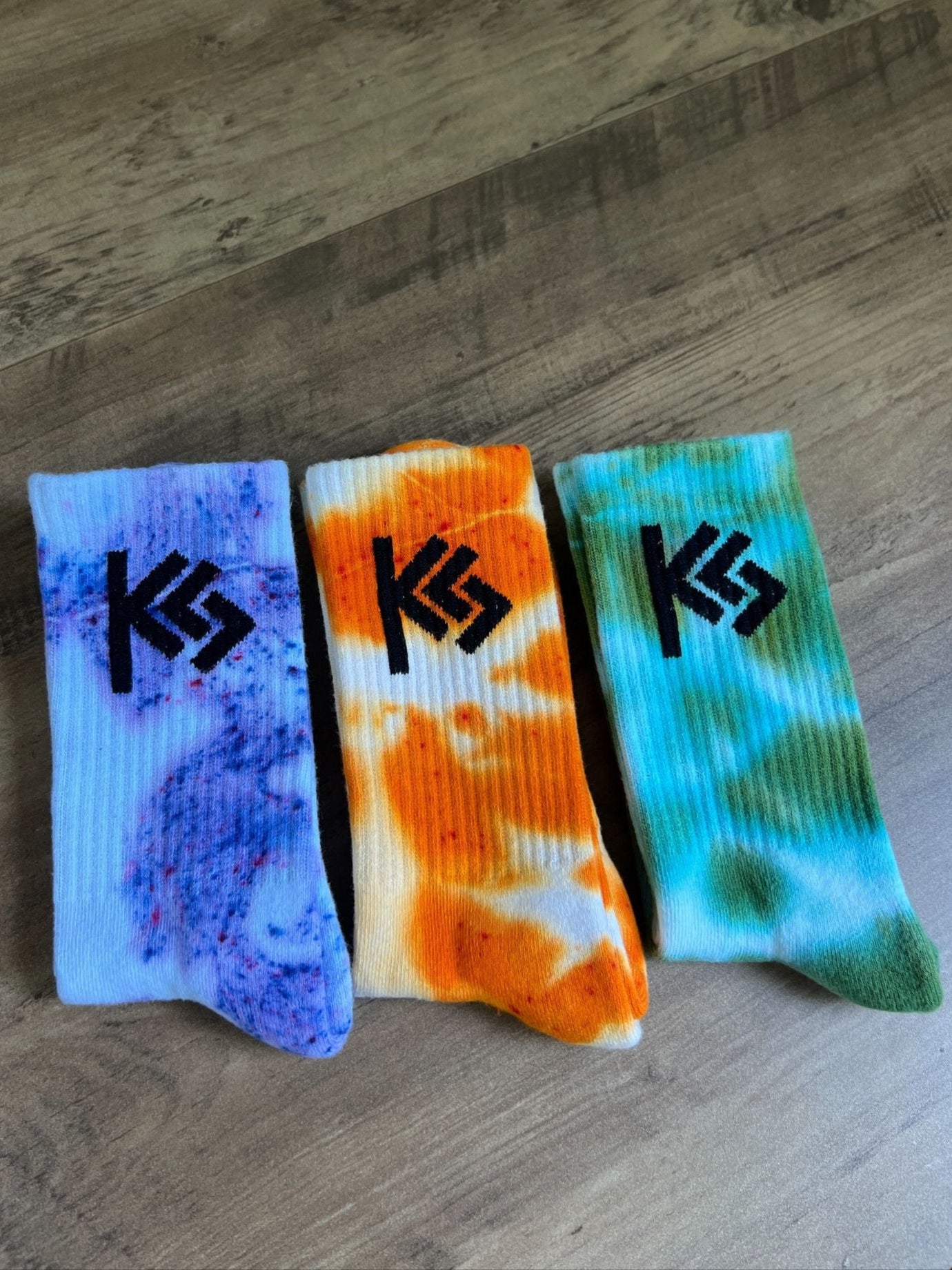 Three colorful socks with tie-dye patterns and a logo on a wooden surface